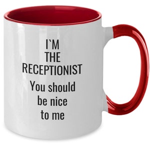 May include: White ceramic mug with a red interior and handle. The mug features the text "I'M THE RECEPTIONIST You should be nice to me" in black, a humorous statement for office settings.