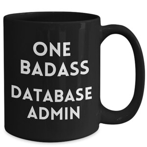 Może przedstawiać: Czarny ceramiczny kubek z białym napisem "ONE BADASS DATABASE ADMIN". Kubek ma zakrzywiony uchwyt i błyszczące wykończenie. Przeznaczony do gorących napojów. Tekst jest pogrubiony, bezszeryfowy.