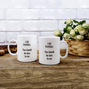 Puede incluir: Dos tazas de cerámica blanca con texto negro que dice "I DO PAYROLL, You should be nice to me." Las tazas están sobre una superficie de madera con un fondo de ladrillo blanco. Una cesta de flores está en el fondo.