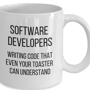 May include: White ceramic mug with the text "SOFTWARE DEVELOPERS WRITING CODE THAT EVEN YOUR TOASTER CAN UNDERSTAND" in black. The mug has a large handle and is designed for hot beverages.