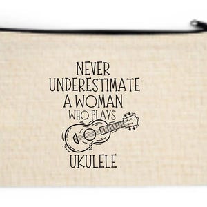 Puede incluir: Bolsa beige con cremallera con el texto negro "NEVER UNDERESTIMATE A WOMAN WHO PLAYS UKULELE". Una ilustración de ukelele en blanco y negro está debajo del texto. La bolsa es rectangular.