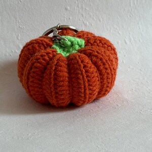 May include: A crocheted pumpkin keychain in shades of orange and green. The pumpkin is round with ribbed detailing and a green stem. A silver-colored keyring is attached to the top. This handmade item is perfect for fall.