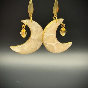 May include: A pair of crescent moon earrings featuring a cream-colored marbled resin finish with gold-toned metal edges. Each earring hangs from a gold-toned hook and includes a small, faceted, translucent amber-colored crystal bead charm.