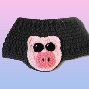 Könnte beinhalten: Ein dunkelgraues gehäkeltes Stirnband mit einem rosa und weißen Schweinegesicht. Das Schweinegesicht hat große schwarze Augen und eine rosa Schnauze. Das Stirnband ist handgefertigt, wahrscheinlich für ein Kind oder Baby.