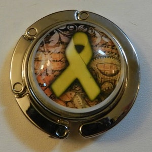 May include: A silver-colored purse hook with a yellow ribbon design. The ribbon is centered on a background of gears and vintage paper. The hook is round with a hinged design for easy storage and use.