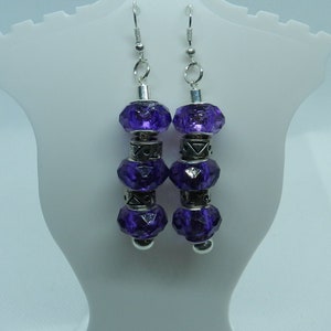 May include: A pair of dangle earrings featuring faceted, deep purple beads. The earrings have silver-colored metal accents and ear wires. The beads are stacked between decorative metal pieces, creating a layered effect. The earrings are displayed on a white stand.