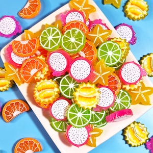 May include: A collection of colorful resin cabochons shaped like fruit slices, including dragon fruit, lime, orange, and star fruit, arranged on a white square plate against a light blue background. The pieces feature vibrant colors and details.