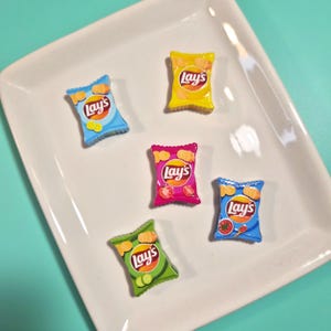 May include: A collection of miniature Lay's potato chip bags in various colors, including yellow, blue, pink, green, and blue. Each bag features the Lay's logo and a depiction of the chip flavor. The bags are arranged on a white rectangular plate.
