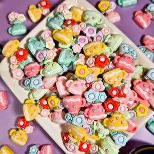 May include: A collection of colorful, miniature teacups and flower decorations in various pastel shades of pink, yellow, green, and blue. The decorations are arranged on a white square plate against a purple background.