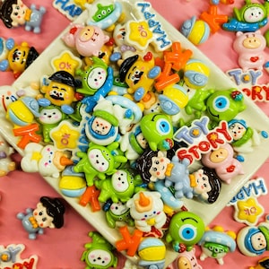 May include: A collection of colorful, cartoon-like embellishments, including characters and shapes, are scattered on a pink surface and a white dish. The embellishments feature the words "HAPPY", "OK", and "TOY STORY".