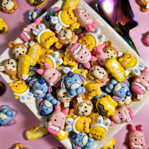 May include: A collection of colorful, cartoon character-themed embellishments. The assortment includes yellow, pink, blue, and brown figures, some with the word "Pooh" on them. The items are arranged on a white dish against a pink background.