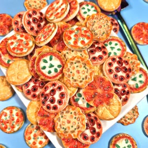 May include: A collection of miniature pizza charms with various toppings, including pepperoni, green herbs, and cheese. The charms are arranged on a white plate against a light blue background. The pizzas are in shades of red, orange, and white.