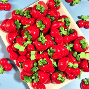 May include: A collection of small, glossy red strawberry-shaped charms with bright green stems, piled together on a light-colored rectangular tray. Each charm features a smooth, rounded surface with a realistic fruit texture and vibrant green leaves.