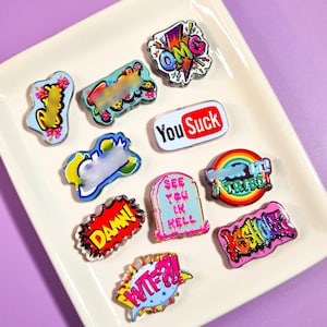 May include: A collection of ten colorful enamel pins featuring various graphic designs and text including OMG, You Suck, Damn, See You In Hell, I Tried, and WTF. The pins display vibrant colors like pink, yellow, red, and blue in comic book styles.