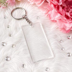May include: A clear rectangular acrylic keychain blank featuring a silver-toned metal split ring attached by a short chain. The transparent pendant has smooth, rounded edges and a flat surface, providing a blank space for customization or design.