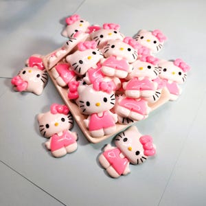 May include: A selection of white and pink Hello Kitty figurines. The figurines have a pink bow, dress, and details. They are arranged on a rectangular, light-coloured tray, with some scattered around it. The background is a light blue surface.