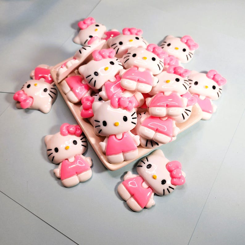 May include: A selection of white and pink Hello Kitty figurines. The figurines have a pink bow, dress, and details. They are arranged on a rectangular, light-coloured tray, with some scattered around it. The background is a light blue surface.