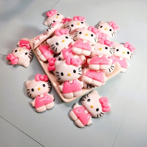 May include: A collection of white and pink Hello Kitty figurines with black whiskers and yellow eyes. The figurines are wearing pink dresses and bows. Some are arranged on a rectangular, off-white tray.