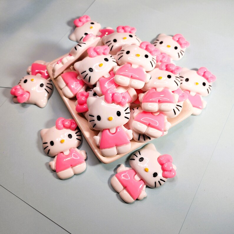 May include: A collection of white and pink Hello Kitty figurines with black whiskers and yellow eyes. The figurines are wearing pink dresses and bows. Some are arranged on a rectangular, off-white tray.