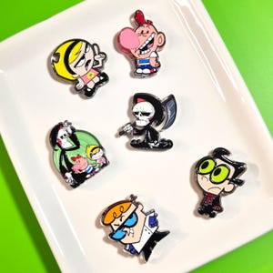 May include: A collection of six metal enamel pins featuring cartoon characters including a blonde girl in a pink shirt, a boy with a red cap and bubblegum, a skeleton with a scythe, a group of three characters, a boy with green eyes, and a boy in a lab coat.