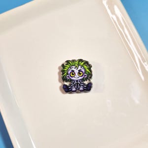 May include: A small pin featuring a cartoon character with bright green hair, large yellow eyes, and pale skin. The character wears a black and white striped outfit with a diamond pattern on the chest and dark purple shoes, sitting in a seated position.