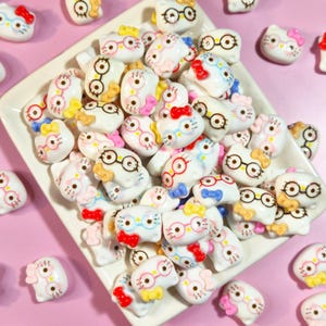 May include: A collection of small, white, cat-shaped resin cabochons featuring printed black-rimmed glasses and colorful bows in red, yellow, blue, and pink. The cat figures are arranged on a white rectangular tray against a light pink background.