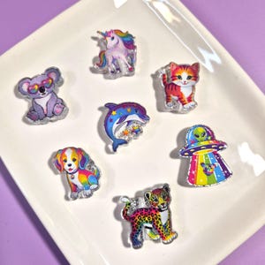 May include: A collection of seven colorful acrylic charms featuring a koala with heart-shaped glasses, a unicorn, a tabby kitten, a dolphin with stars, a rainbow-colored puppy, a leopard, and an alien in a flying saucer with a rainbow beam.