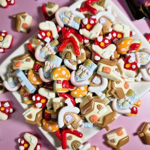 May include: A collection of miniature, colorful mushroom and house-shaped embellishments. The assortment includes red, orange, yellow, and brown houses and mushrooms with white polka dots. The items are arranged on a white plate against a pink background.