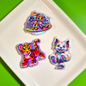 May include: Three acrylic flatback charms featuring vibrant, multi-colored illustrations. One charm depicts two aliens on a flying saucer, another shows a tiger cub with orange and black stripes, and the third is a fluffy cat with rainbow fur.