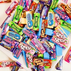 May include: A collection of colorful, miniature gum wrappers with various brand names and flavors, including Doublemint, Razzles, Hubba Bubba, Jolly Rancher, and Big Ouch! The wrappers are arranged in a random pattern on a white surface.