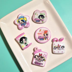 May include: Six flat resin charms featuring The Powerpuff Girls characters. Shapes include a star with Bubbles, a cat head with Buttercup, a rectangle with Buttercup, a circle with Blossom, a square with bunny ears, and a bottle shape with all three.