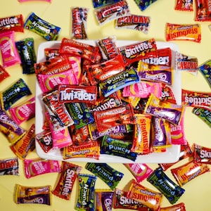 Puede incluir: Un plato blanco lleno de una variedad de envoltorios de caramelos coloridos. Los envoltorios presentan marcas populares de caramelos como Skittles, Starburst, 100 Grand y Twizzlers.