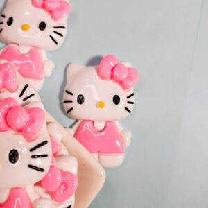 May include: Close-up of several white and pink Hello Kitty figurines. Each figurine has a pink bow, a pink dress, and black eyes and whiskers. The background is a light blue colour.