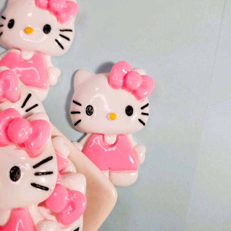 May include: Close-up of several white and pink Hello Kitty figurines. Each figurine has a pink bow, a pink dress, and black eyes and whiskers. The background is a light blue colour.