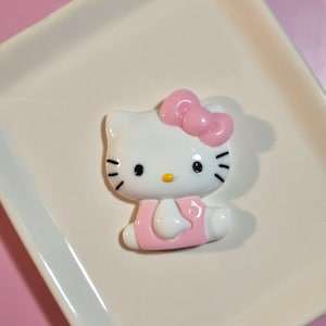 May include: A white Hello Kitty character figurine with a pink bow on the head and a pink outfit. The character has black eyes, black whiskers, and a small yellow nose. The figurine is made of a smooth, glossy material and is shaped like a flat charm.