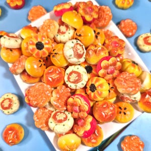 May include: A collection of colorful, decorative candies in various shapes, including flowers and pumpkins, arranged on a white plate. The candies are primarily orange, yellow, and red, with some green and white accents. The candies are likely handmade and suitable for a party.