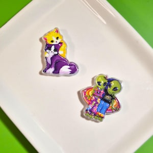 May include: Two acrylic pins featuring colorful illustrations. One pin depicts a yellow kitten hugging a purple and white cat. The second pin shows two green aliens with large black eyes, one wearing a floral dress and the other a black shirt.