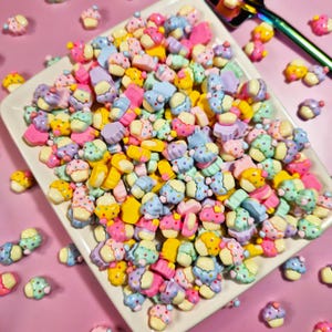 May include: A collection of colorful miniature cupcake-shaped charms in various pastel colors, including pink, blue, yellow, and purple. The charms are arranged on a white rectangular plate, perfect for crafting or jewelry making.