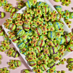 Puede incluir: Una colección de pequeñas figuras de tortugas verdes con máscaras de colores, incluyendo rojo, azul, naranja y morado. Las figuras están dispuestas sobre un plato blanco y dispersas sobre una superficie rosa. Las tortugas tienen detalles y son de colores brillantes.