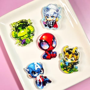 May include: Five acrylic pins featuring chibi-style illustrations of Marvel characters: a green Hulk, a silver-helmeted Thor, a red and blue Spider-Man with galaxy-patterned eyes, a green and gold Loki, and a blue Captain America with a shield.