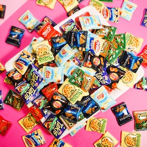 May include: A white plate filled with miniature bags of various popular snack brands, including Lay's, Doritos, Cheetos, Fritos, and Takis. The bags are colorful and feature the brand logos and product names.
