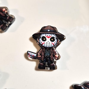 Puede incluir: Un pequeño pin de acrílico con un personaje de dibujos animados que lleva un sombrero fedora marrón y una máscara de hockey blanca con marcas rojas. El personaje viste un abrigo marrón y sostiene un cuchillo plateado con manchas rojas.