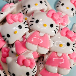 May include: A selection of white and pink Hello Kitty character charms. The charms depict the famous cat with a pink bow, black whiskers, and a yellow nose. Some charms are wearing pink outfits. These charms are suitable for crafting.
