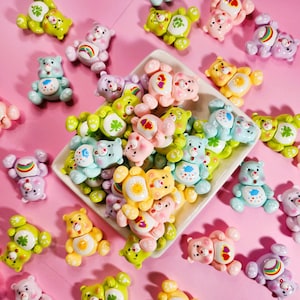 May include: A collection of colorful, small, plush bear figurines with various designs and patterns. The bears are arranged on a pink background.