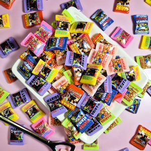 May include: A collection of colorful HARIBO candy packages in various sizes and designs, scattered on a white tray and a pink surface. The packages feature text and graphics, with a pair of scissors in the lower left corner.