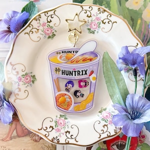 Puede incluir: Un charm de acrílico transparente con forma de cuenco de ramen, con el texto "HUNTRIX" e ilustraciones de personajes. El charm cuelga de un cierre dorado en forma de estrella. El charm está sobre un plato decorativo con detalles florales y rodeado de flores moradas.
