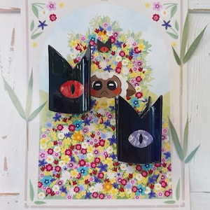 May include: Two black, crown-shaped brooches with red and purple eye details are displayed in front of a floral illustration. The illustration features a lamb with a crown and a colorful flower background. The brooches are likely accessories.