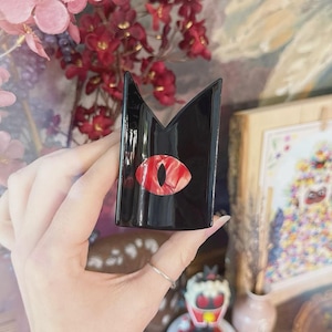May include: A black, glossy decorative object with a stylized red and white eye design. The object has a unique, angular shape and is held in a hand. The background features flowers and artwork.