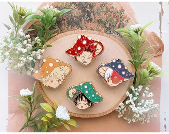 May include: Four enamel pins featuring anime characters with mushroom caps. The pins have gold-colored borders and are in various colors: red, orange, blue, and green. The pins are arranged on a wooden slice with floral accents.