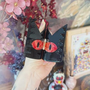 May include: Black hair clip with a crown design and red eye accents. The clip features a unique design with a central clasp and decorative elements. A stylish hair accessory for various hairstyles.